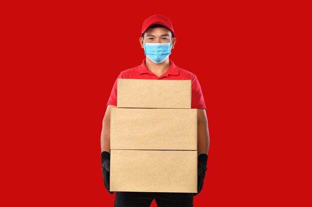 Happy young asian delivery man in red uniform, medical face mask, protective gloves carry cardboard box in hands on red background. delivery guy give parcel shipment. during covid-19 outbreak