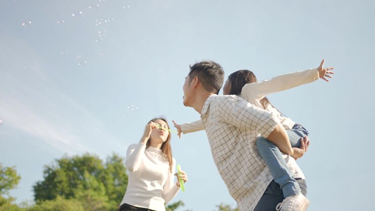 Photo 幸せな若いアジア人の父親が小さな女の子とおんぶし、母親が公園で彼女と遊ぶために走って、緑豊かな庭園で一緒に楽しんだ自然の中で晴れた秋の日を楽しむ家族