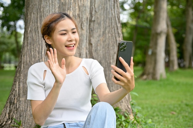 Happy young Asian female relaxes while sitting under the tree using her smartphone