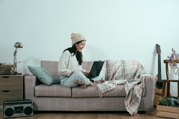 幸せな若いアジアの韓国人女性は、ソファに足を置いてリラックスしてラップトップコンピューターで電子書籍をお楽しみください。コピースペースのある側面図。ノートパソコンでオンラインショッピングソファでセーターとボブル帽子の屈託のない女の子