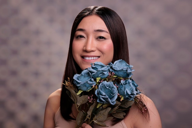 Happy young asian lady with blue roses present portrait. Smiling cheerful natural beautiful fashion model posing with blooming flowers bouquet in hands for studio photoshoot