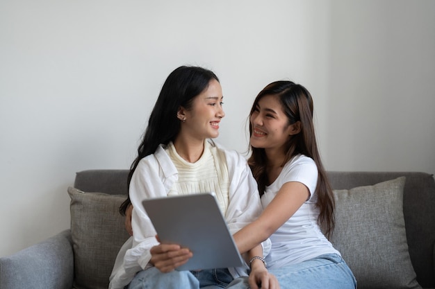 Happy young asian lesbian couple hugging having fun using digital tablet relaxing on couch at home Two smiling women friends holding computer looking at screen enjoying surfing online watching video