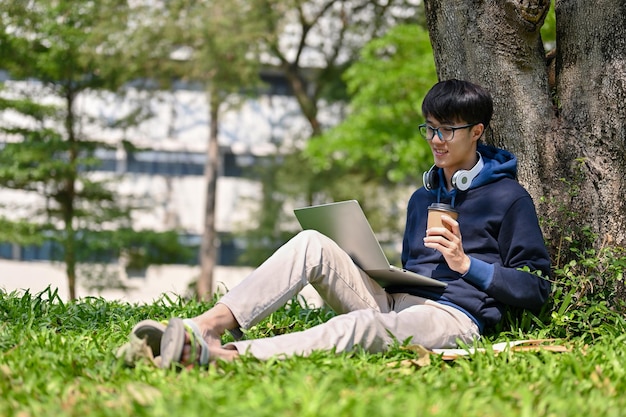 Happy young Asian male college student using his laptop under the tree in the campus park