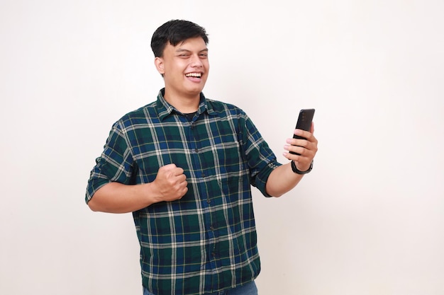 Счастливый молодой азиат сжимает руку, держа в руках сотовый телефон.