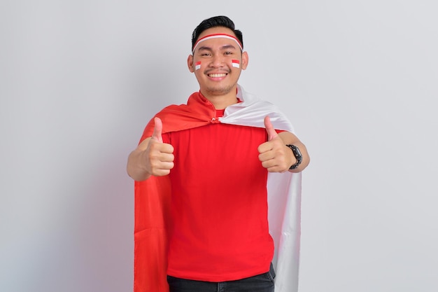 Happy young Asian man holding the Indonesian flag while showing thumbs up isolated on white background Indonesian independence day concept