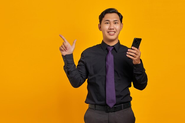 Happy young Asian man holding mobile phone and pointing finger to copy space on yellow background