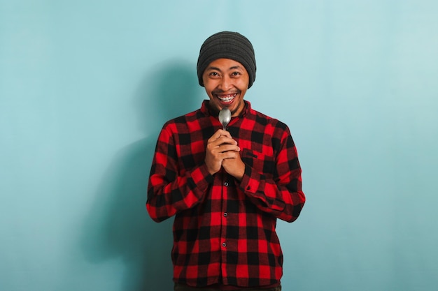 Happy Young Asian man holding spoon near his mouth feeling excited to eat isolated on blue backdrop