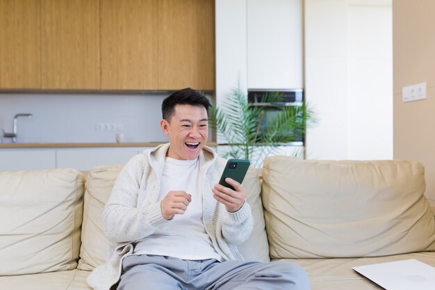 happy young asian man at home looking at cellphone with emotion of winner