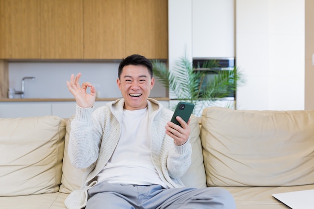 집에 있는 행복한 젊은 아시아 남자가 승자의 감정으로 휴대폰을 보고 있다
