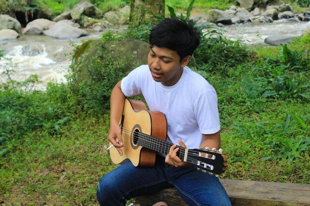 Happy young asian man is playing acoustic guitar and singing in the riverside when picnic on holiday