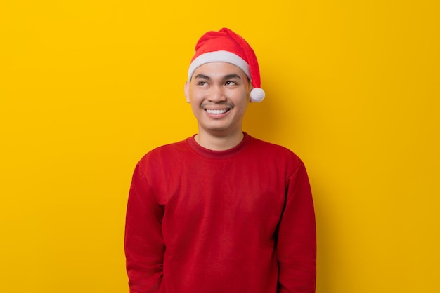 Happy young asian man in santa hat looking upwards with broad smile on yellow studio background celebration christmas holiday and new year concept