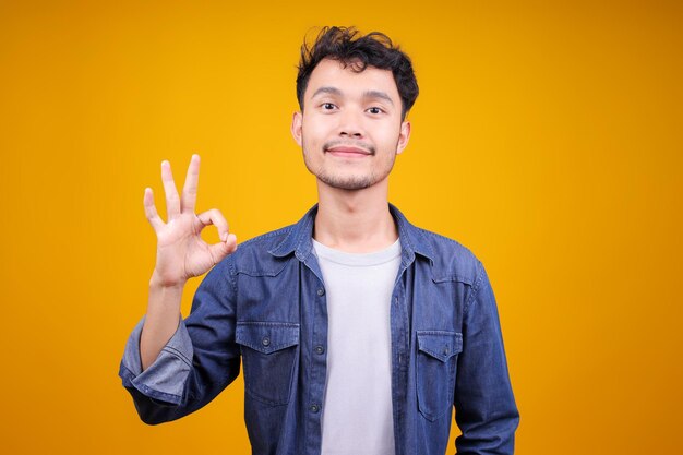 黄色いスタジオの背景でカメラにOKジェスチャーを示す幸せな若いアジア人男性