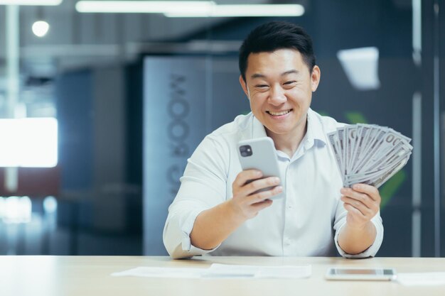 A happy young asian man sits in the office at the table holds a\
phone and cash money in his hand a