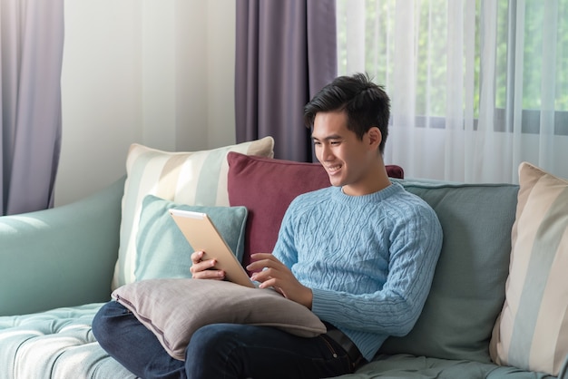 Happy young asian man sitting on the sofa watching a tablet at home.