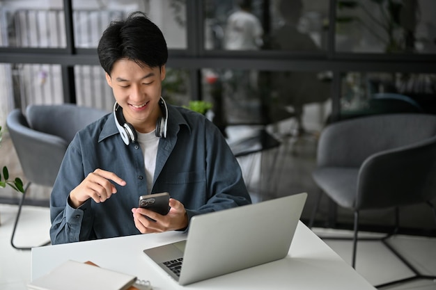 커피숍에서 원격으로 일하는 동안 행복한 젊은 아시아 남자가 휴대폰으로 문자를 보고 웃고 있다