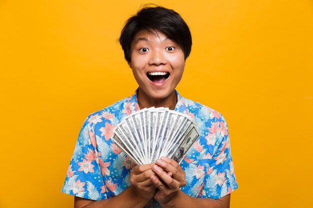 Happy young asian man standing isolated over yellow space holding money