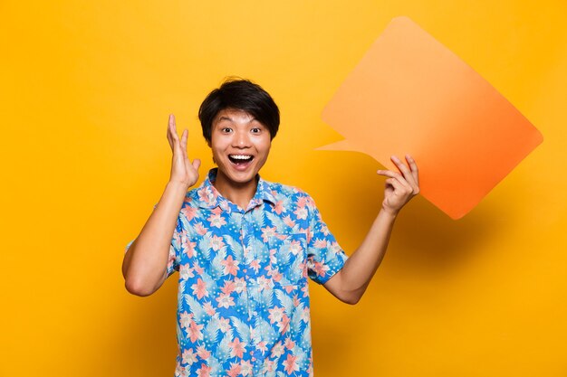 Happy young asian man standing isolated over yellow space holding speech bubble.