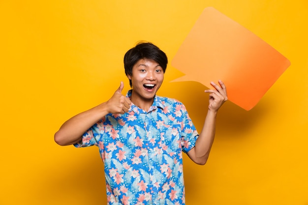 Happy young asian man standing isolated over yellow space holding speech bubble.