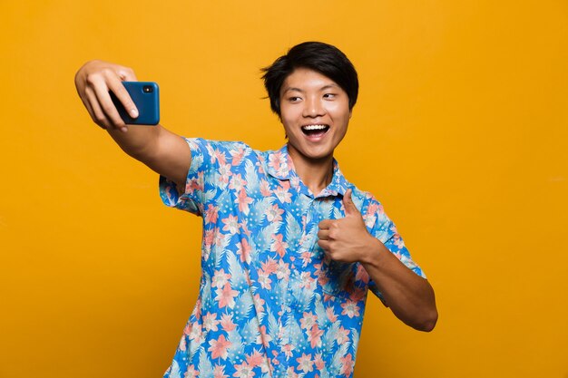 Happy young asian man standing isolated over yellow space take a selfie by mobile phone with thumbs up.