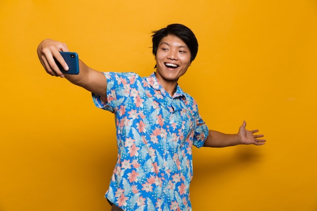 Happy young asian man standing isolated over yellow space take a selfie by mobile phone