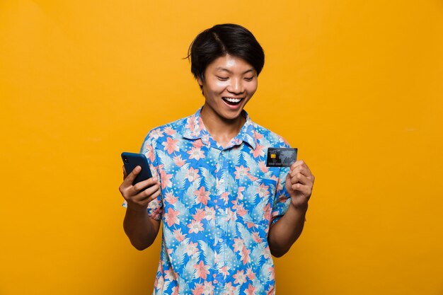 Happy young asian man standing isolated over yellow space using mobile phone holding credit card.