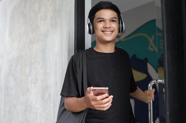 Happy young asian man wearing wireless headphones smiling while opening glass door