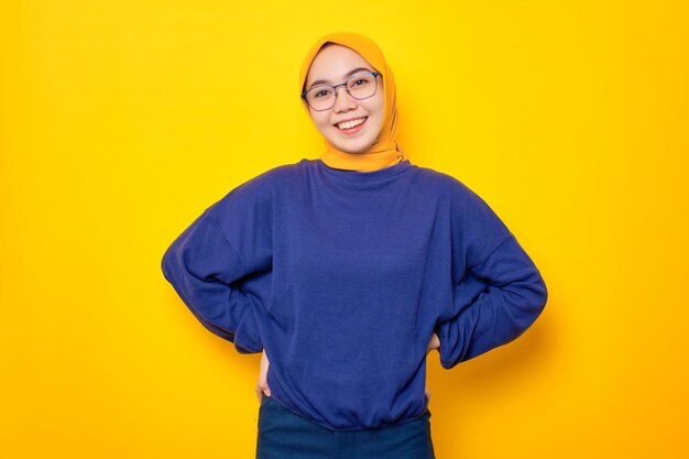 Happy young asian muslim woman dressed in casual sweater holding hands on waist and looking confident isolated over yellow background