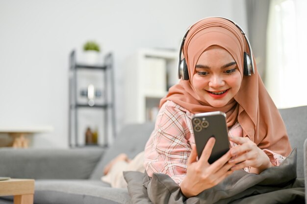 Happy young Asian Muslim woman listening to music through headphones laying on sofa