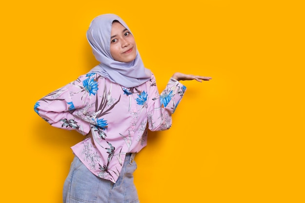 黄色の背景に指でさまざまな方向を指している幸せな若いアジアのイスラム教徒の女性
