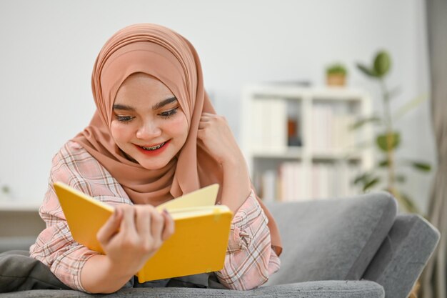 幸せな若いアジアのイスラム教徒の女性が本を読んで、リビング ルームのソファに横になっています。