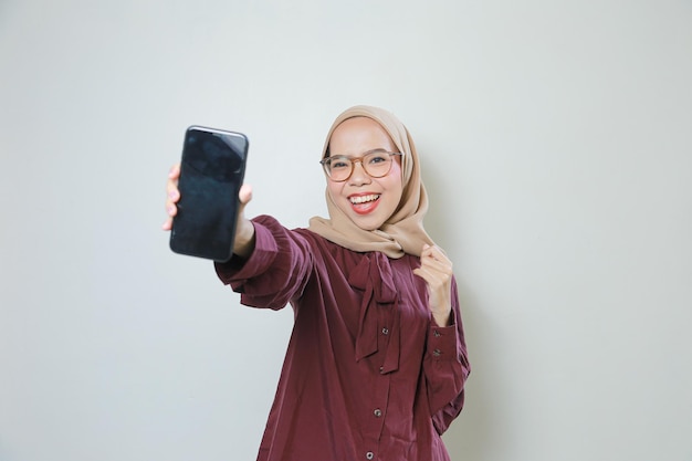 Happy young Asian Muslim woman wearing glasses showing mobile phone to camera isolated