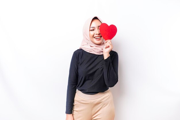 A happy young Asian Muslim woman wearing a hijab feels romantic shapes heart gesture expressing tender feelings and holding a red heartshaped paper