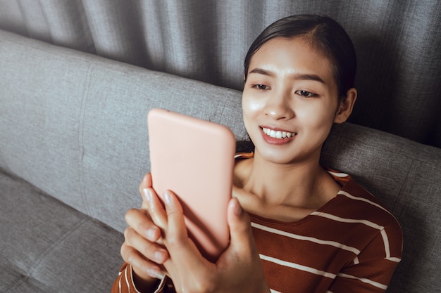 Felice giovane donna asiatica piacevole tenendo lo smartphone in mano a casa, sorridendo e guardando video divertenti.