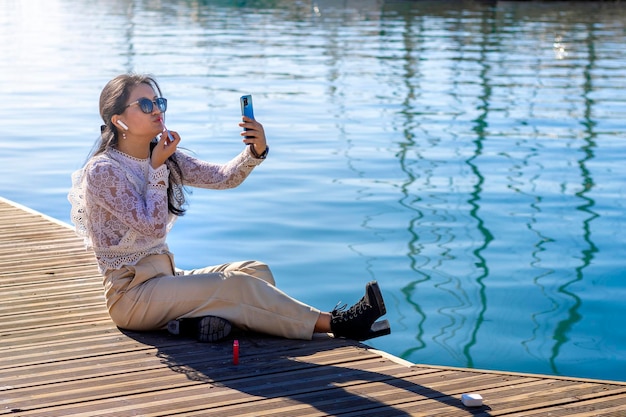 幸せな若いアジアのセールスウーマンは、携帯電話を使用して、港に座っている彼女の唇をペイントします。
