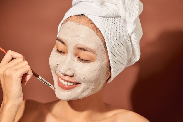 Happy young Asian woman applying clay mask on her face over orange background. Close up.