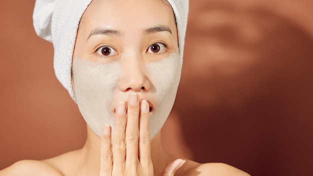 Happy young Asian woman applying clay mask on her face over orange background Close up