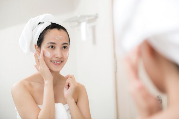 수건을 착용하고 욕실에서 얼굴을 만지는 동안 얼굴 로션을 바르는 행복한 젊은 아시아 여성
