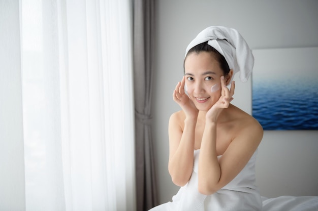 Happy young Asian woman applying face lotions while wearing a towel and touching her face Daily makeup and skincare