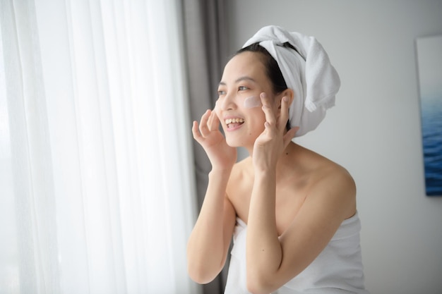 Happy young Asian woman applying face lotions while wearing a towel and touching her face Daily makeup and skincare