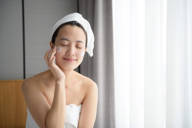 Happy young Asian woman applying face lotions while wearing a towel and touching her face Daily makeup and skincare