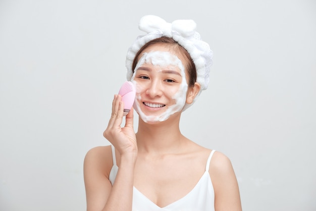 Happy young Asian woman applying foaming cleanser on her face and wearing towel on her head.