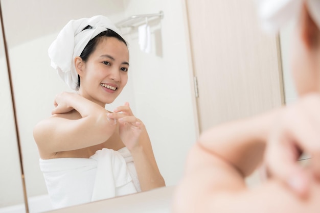 Счастливая молодая азиатка наносит лосьоны на тело в ванной комнате
