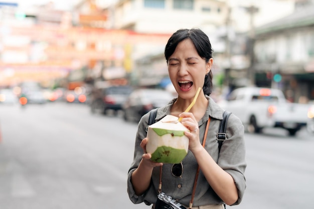 幸せな若いアジア女性のバックパック旅行者は、タイのバンコクの中華街の屋台市場でココナッツ ジュースを飲みながら、脇道をチェックする旅行者