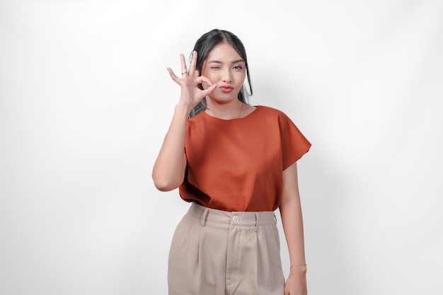 茶色のシャツとタウペのズボンを着た幸せな若いアジア人女性が白い背景に隔離されたOKの良い手のジェスチャーをしています
