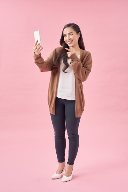 Happy young asian woman casually dressed standing isolated over pink background, using mobile phone