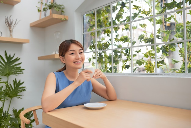 집에서 창문 뒤에서 커피를 마시는 행복한 젊은 아시아 여성