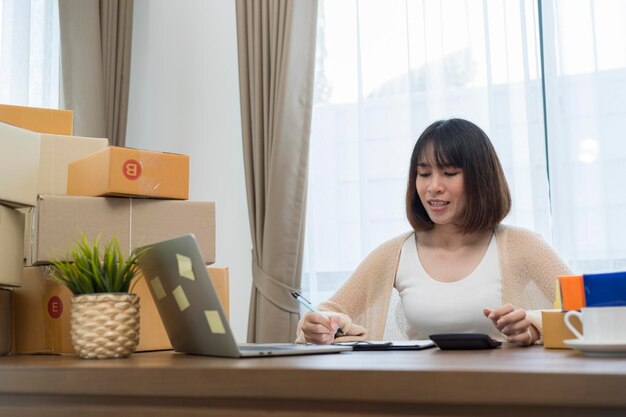 Счастливая молодая азиатская женщина-предприниматель улыбается за успех продаж после проверки заказа в интернет-магазине на ноутбуке дома
