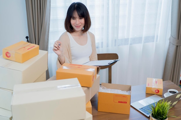 Happy young Asian woman entrepreneur Smile for sales success after checking order from online shopping store in a smartphone at home office