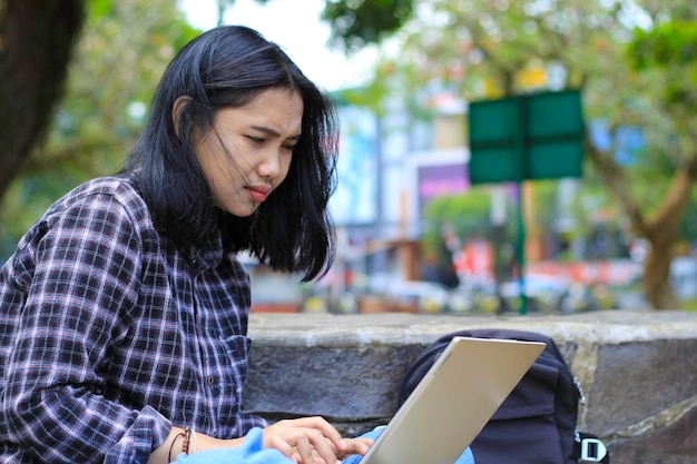 幸せなアジアの若い女性はラップトップを使ってリモートで作業し,屋外でソーシャルメディアを ⁇ 覧することに集中しています