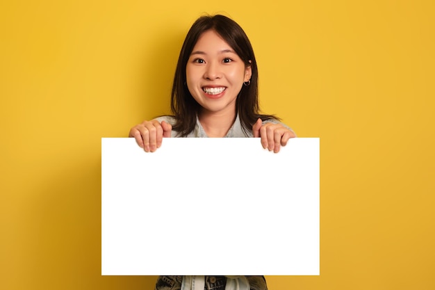 Happy young asian woman holding blank paper banner smiling at camera on yellow studio background